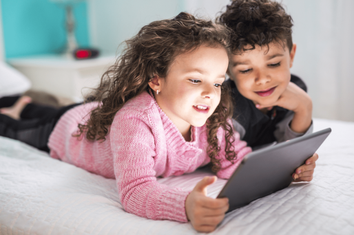 Image of two young children laying on a bed, looking at a tablet.