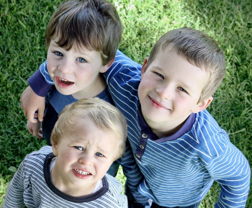 Three children standing outside on grass looking up at the camera and smiling.